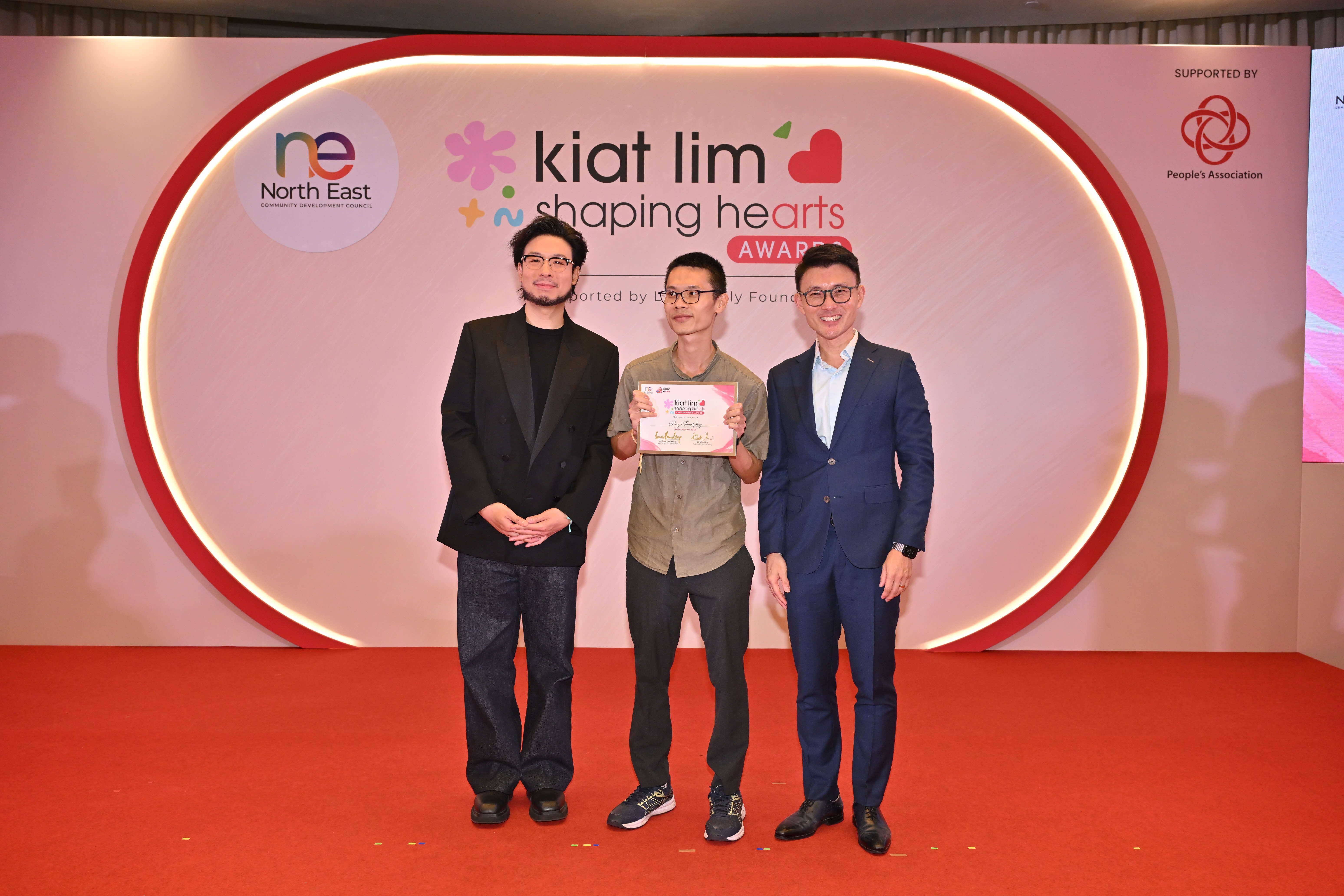 Award recipient standing on stage with the presenter, holding their certificate and posing for a group photo during the award ceremony, with a backdrop reading ‘Kiat Lim-Shaping Hearts Awards'.
